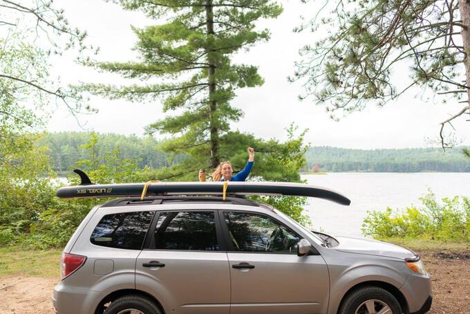 person waving by vehicle, forest and lake in background